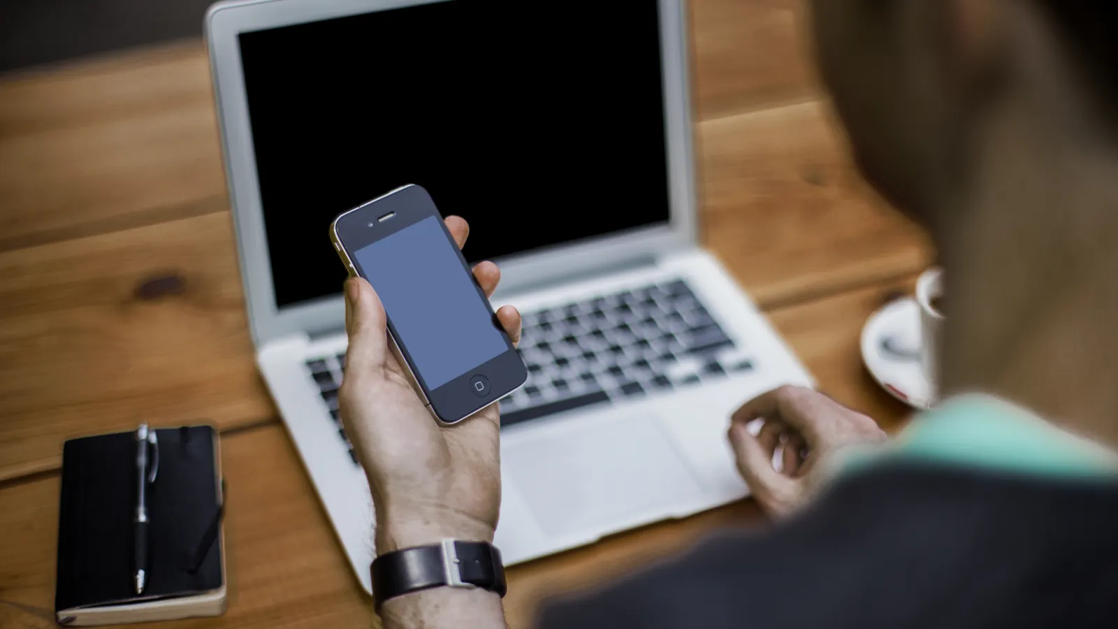 A hand holding a smartphone over a laptop on a wooden desk